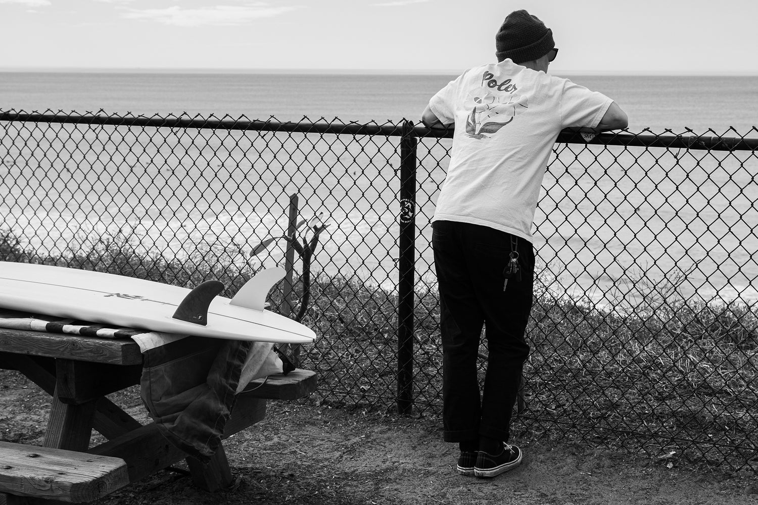 A man leaning against a fence looking out to sea with his surfboard on a picnic table.