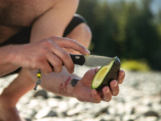A man slicing an avocado by the river with his Carter EDC pocket knife.