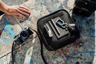 A camera next to a bag with a knife, pen, and carabiner on top.